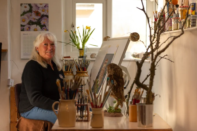 Eleanor Ludgate, artist. Eleanor is sat at her easel facing the camera, surrounding her are various pots of paint brushes, a stuffed owl and a small tree. on the easel is the painting of the owl Eleanor is completing.