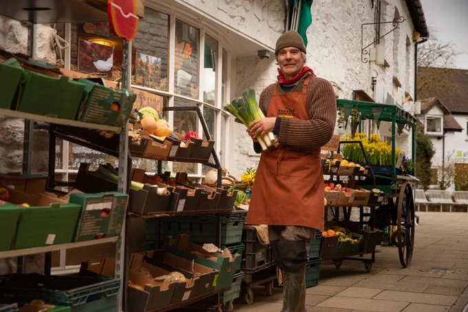 Hilton Organic cooperative. Gentleman standing outside the shop holding leeks. Along the front of the shop are various grocer boxes on shelves containing fruit and vegetables, along with a small cart holding flowers. 