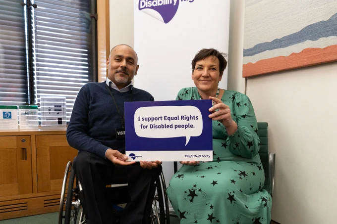 This is an image of Kamran Malik, the CEO of Disability Rights UK and Tonia Antoniazzi MP holding a sign that states "I support Equal Rights for Disabled People". Kamran is a wheelchair user, wearing a navy jumper and black trousers. Tonia is sat besides him wearing a green dress with dark stars. 