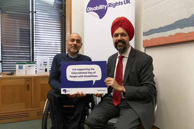 This is an image of Kamran Malik and Tan Desi sitting beside each other and holding a sign that reads "I'm supporting the International Day of People with Disabilities". There is a white banner in the background with a purple Disability Rights UK logo. 