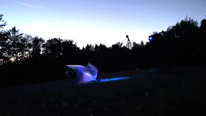 A photography by Mo Costello. A microphone and lights are set up in a field, with trees along the horizon line set against a white and blue sky.