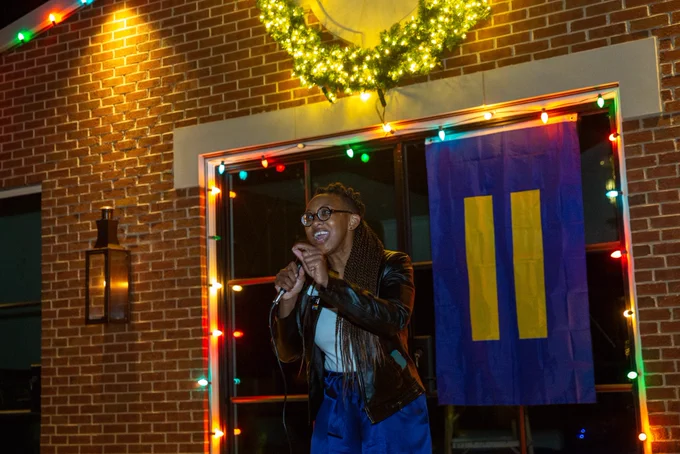 Kelley, standing in front of an HRC flag attached to a home adorned with Christmas lights, speaks passionately into a microphone to an off camera audience.