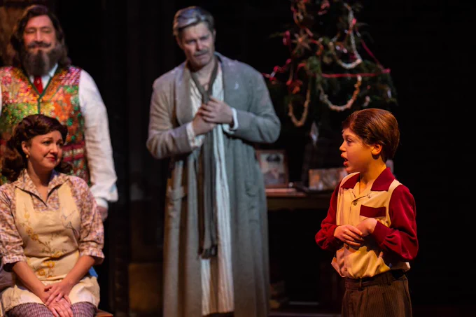[Image Description: Dressed in 1950's era attire, a boy, Tim, stands to the right singing, while a woman, his mother, Emily, sits listening to him. Behind her are two men, the Ghost of Christmas Present and Ebenezer Scrooge, also watching the boy. A Christmas tree is seen in the background.]