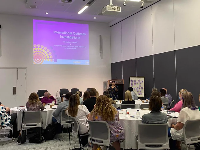 The photo shows several people seated on chairs, listening to a woman speaking. The slide on the front reads: International Outbreak Investigations. 

The photo was taken during the Australasian Infection Prevention and Control International Outbreak Conference in 2022.