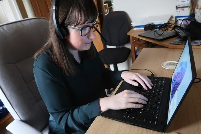 Holly sat at a desk wearing a headset and typing on a laptop. She's wearing a green blouse.