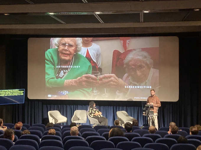 A man stands on stage at a podium in front of an audience with a woman sitting on a seat to his left. He is wearing a brown top and cream trousers and is in front of a screen featuring two elderly women clinking wine glasses