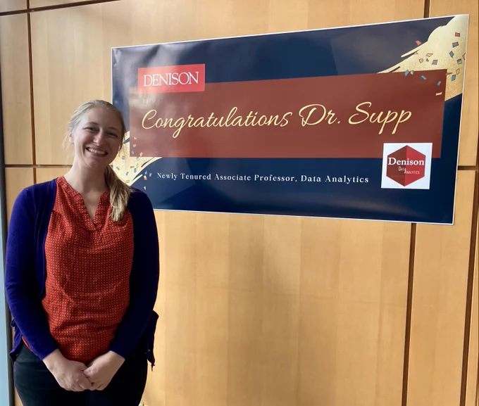 Photo shows a smiling woman with blonde hair named Dr. Supp next to a large sign that reads "Congratulations Dr. Supp. Newly tenured Associate Professor of Data Analytics at Denison University".