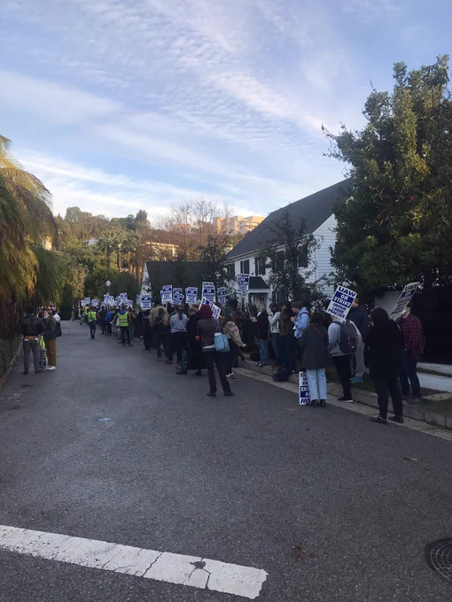 A large crowd of striking academic workers outside an outdated Federal-Revival style mansion in which resides UC Regent Jay Sures in Brentwood, Los Angeles. The crisp dawn light illuminates the background foliage.