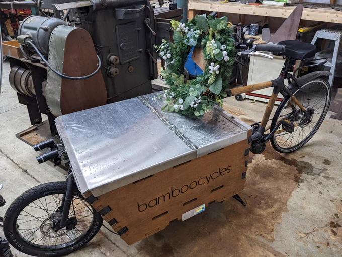 A bamboo cargo bike, sitting in a rather cluttered shop, with a large holiday wreath hanging from the handlebars.