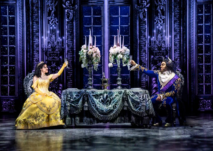 Belle (in a yellow dress) and Beast (in a blue suit) sit at opposite ends of a grand dining table, raising a glass of champagne