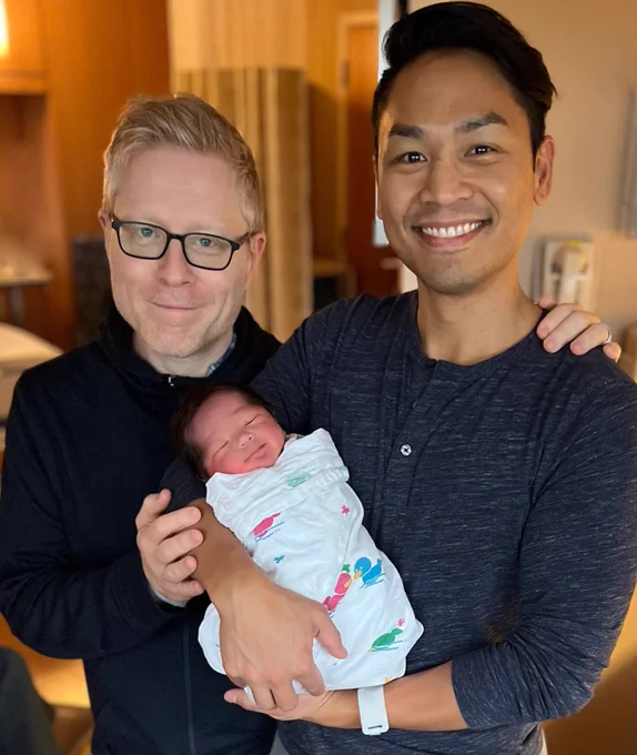 Two smiling men hold a swaddled baby.