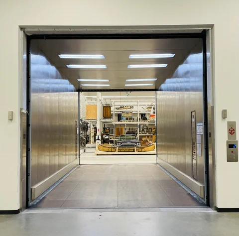 Looking through a massive freight elevator with metallic walls and rectangular lights on the ceiling. On the other side is a canoe, large black car and shelves of artifacts including bicycles, large wooden cabinets and black metal somethings