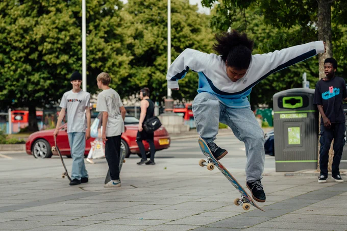young person skateboard flipping outside, other young people stand behind chatting