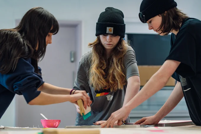 three young people, two of whom are wearing beanie hats, are doing screen printing