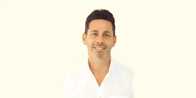 A white man with dark short hair, a dark beard, and blue eyes smiles into the camera. He wears a white shirt and stands in front of a cream colored background.