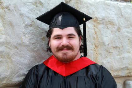 A man poses for a photo. He is smiling while wearing graduation regalia, including a graduation cap with black tassle, and a red stole