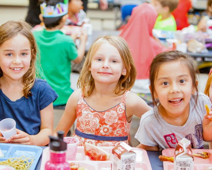 Three smiling students sit at a lunch table in the school cafeteria, their meal trays in front of them.