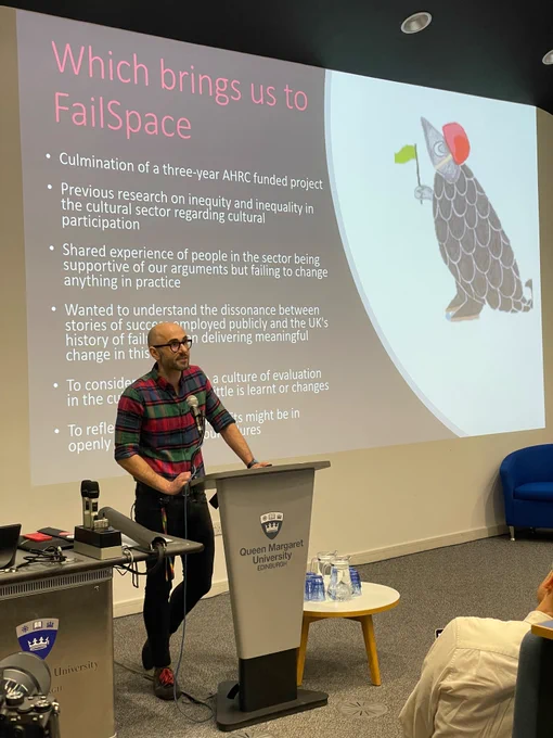 A white man, in black rimmed glasses stands at a lectern speaking energetically in front of a projector that includes statements about failure.