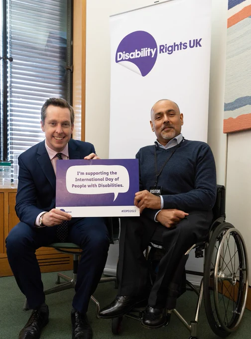 The Minister for Disabled People is seated next to a man in a wheelchair. They are holding a card stating, "I'm supporting the International Day for People with Disabilities." between them.