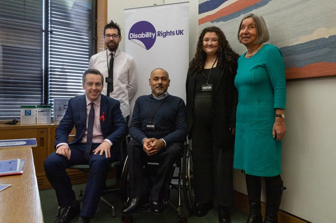 A group photo in an office, with the Minister for Disabled People seated on the left, beside a man in a wheelchair. Three others are standing behind and beside them. A Disability Rights UK banner is visible in the background.