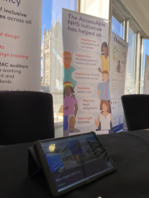 AccessAble website displaying on a tablet, in the background is an AccessAble banner stand listing AccessAble’s services. Outside the window is Tower Bridge, London