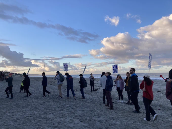 A crowd of workers holding 'UAW on strike' picket signs standing on the beach at sunrise.