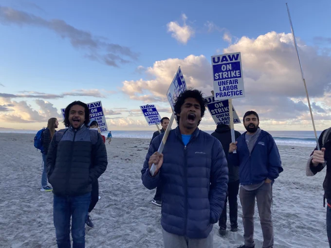 A crowd of workers holding 'UAW on strike' picket signs standing on the beach at sunrise.