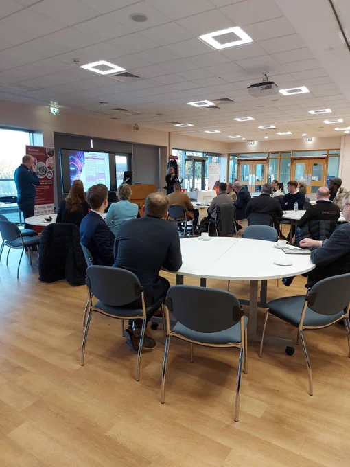 A group of people in a conference room, sat at tables whilst a speaker is at the front.
