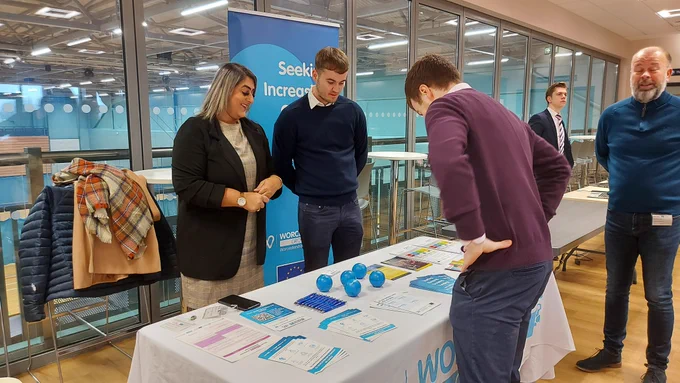 A male talking to a female and male exhibitor, who are stood behind a table, at a business event.