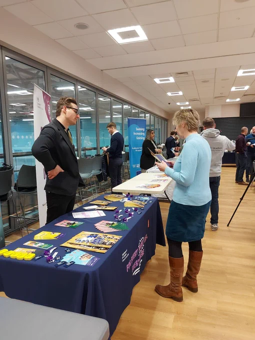 A female talking to a male exhibitor, who is stood behind a table, at a business event.