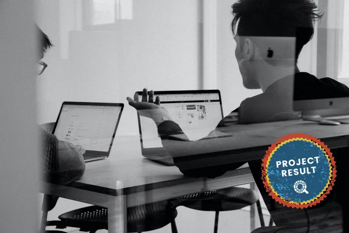 a meeting room, 2 young males sitting around the table with their laptops open and discussing