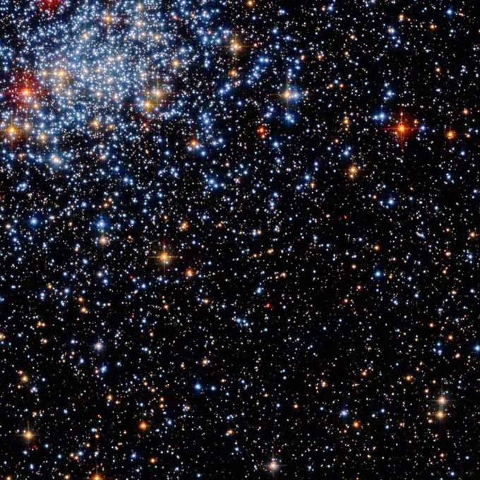 Thousands of stars shine against a black backdrop of space in shades of white, orange, and blue. At the top left of the image, there is a dense cluster of bright, blue stars.