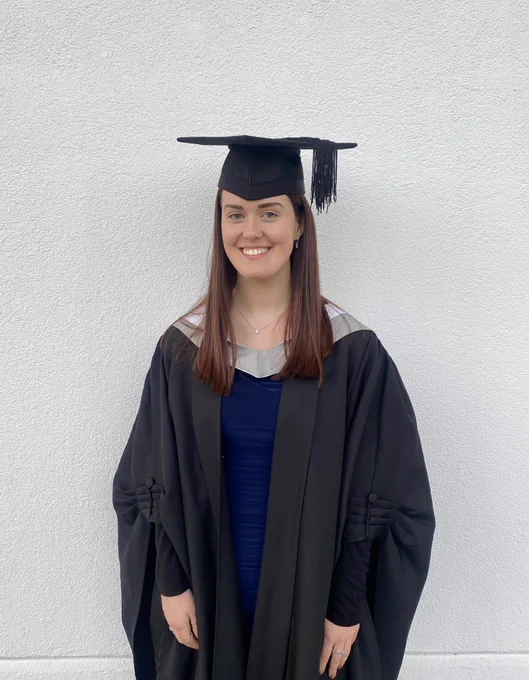 Me smiling in a graduation gown and hat. Underneath my gown I am wearing a blue dress.