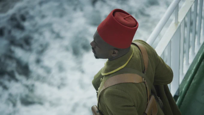 An African soldier seen in three quarter profile from above, in khaki uniform and red fez; looking out to the sea from the deck of a ship.
