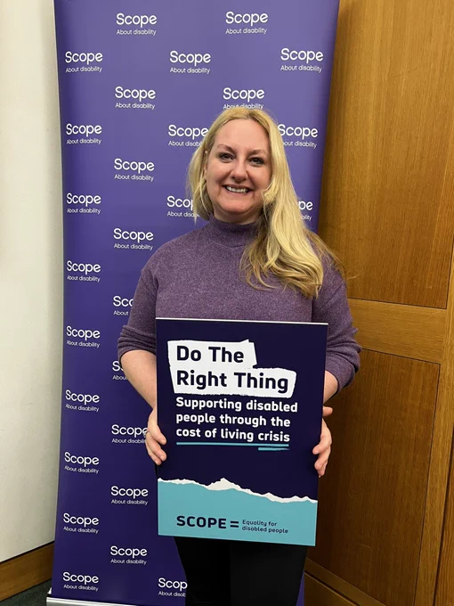 This is an image of Dr Lisa Cameron MP standing in front of a purple banner that says Scope about disability repeatedly. Lisa is holding a sign that says “do the right thing: support disabled people through the cost of living crisis”.