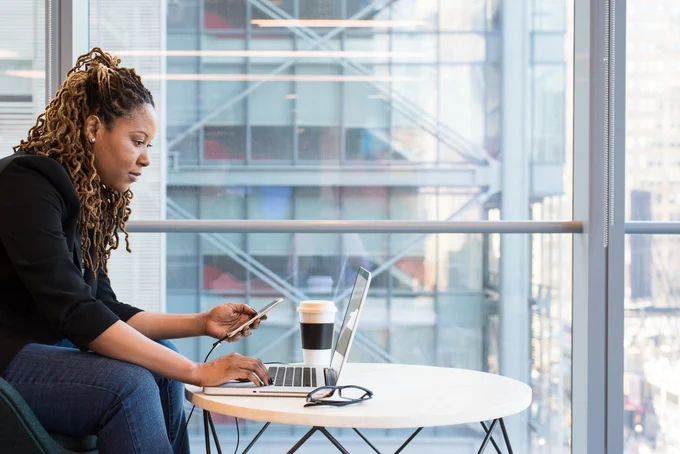 A person with long hair in twists and brown skin sits at a cafe table, using a laptop and cell phone. Also on the table are glasses and a takeout cup of coffee.