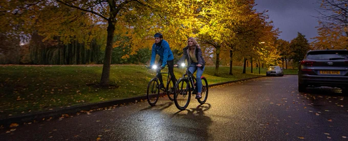 A man and a woman riding bikes side by side on a dark road under streetlights. They both have bright lights on the their bikes. Photo via Cycling UK