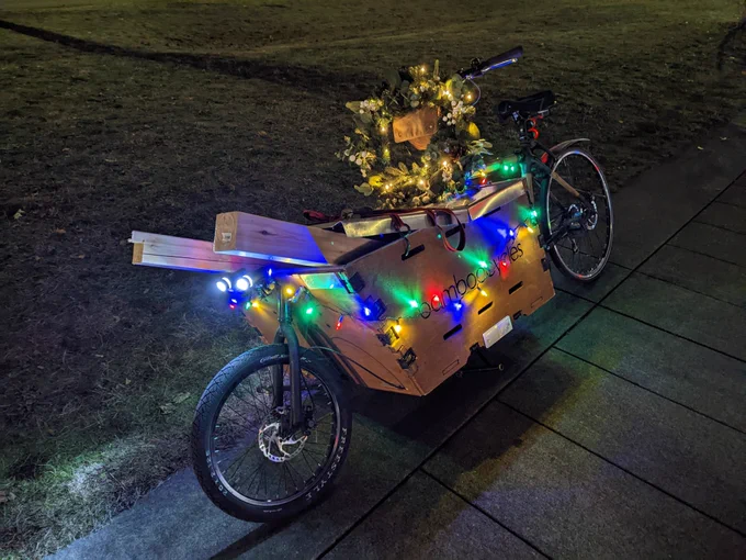 A bakfiets cargo bike at night, festooned with holiday lights and a wreath, with 2 4' sections of 4x4 sticking out of the box.