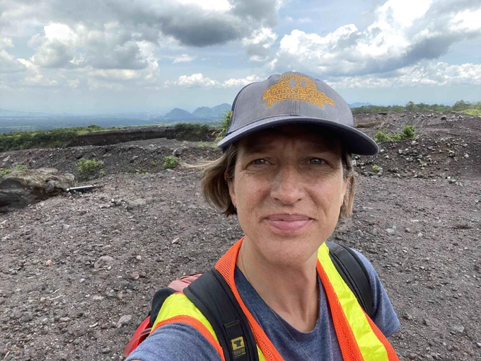 selfie of women in her mid-40s in a volcanic landscape with a baseball cap, daypack, and safety vest on.