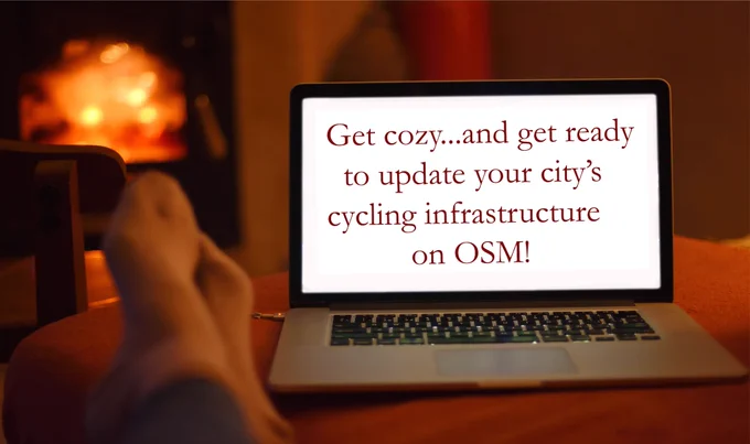 A pair of feet up on an ottoman in front of a fire and a laptop. Laptop screen says "Get cozy...and get ready to update your city's cycling infrastructure on OSM!"