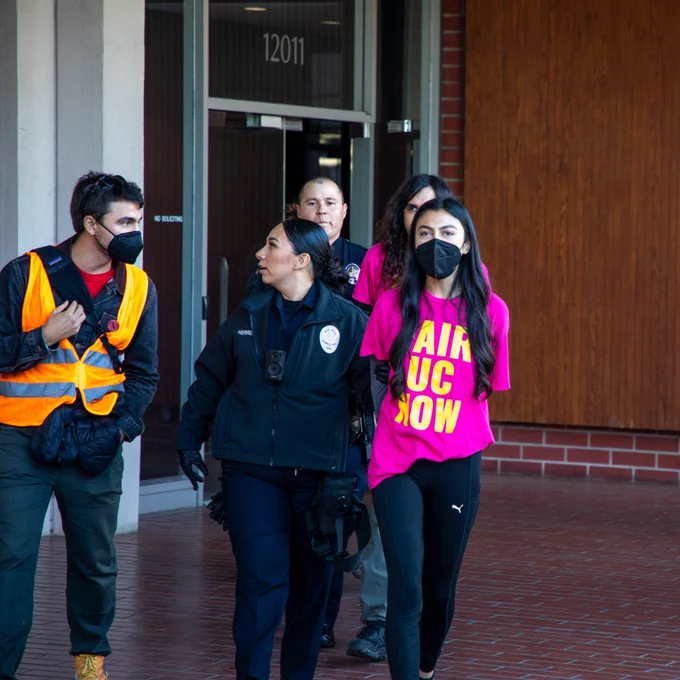 Photo of two workers in pink shirts that say "Fair UC now" are led out of a brick building by cops. 