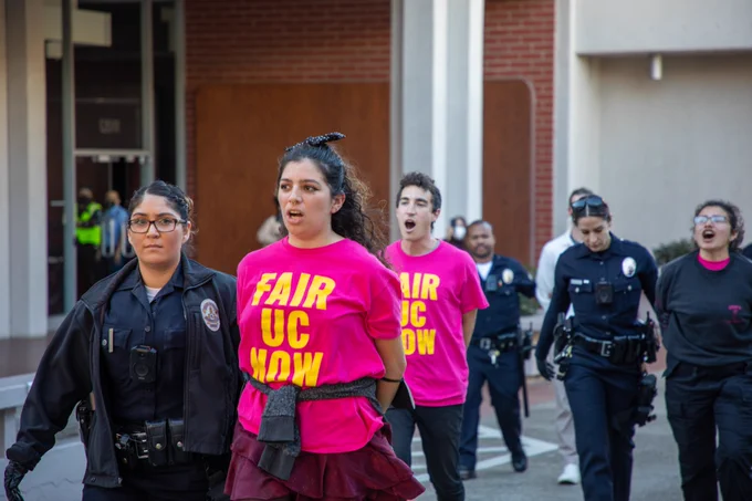 Photo of three workers in pink shirts that say "Fair UC now" are led out of a brick building by cops. 
