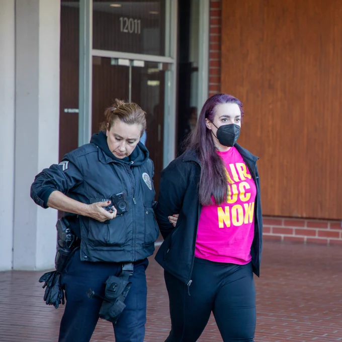 Photo of a workers in a pink shirts that says "Fair UC now" are led out of a brick building by cops. 