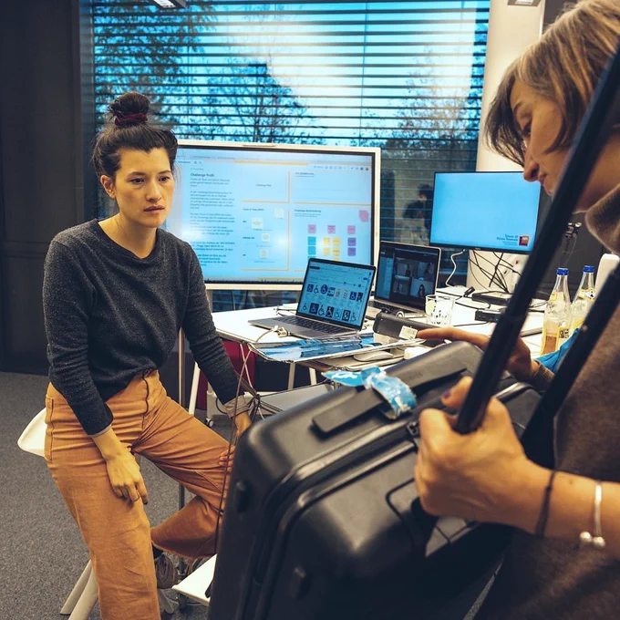 Eine Person hält einen Rollkoffer. Eine weitere Person schaut auf den Koffer. Im Hintergrund mehrere Computerbildschirme.