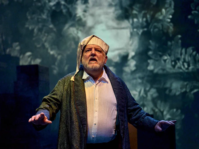 1 - Simon Russell Beale looks upwards with his hands on either side of his face, he wears a nightcap and gown.