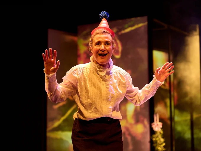 2 - Lyndsey Marshal wears a colourful cardboard cone shaped party hat. Her arms are open and she's grinning.