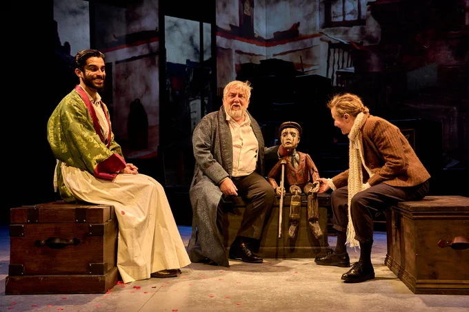 4 - On three wooden chests sit Eben Figueiredo in a long white apron, Simon Russell Beale in an old fashioned dressing gown and sat with a small wooden puppet of a little boy, and Lyndsey Marshal who holds the puppets other hand.