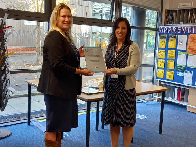 Cllr Chadd and Debbie Mahoney looking at the camera as Cllr Chadd hands over the certificate, standing in a college room with shelves and files, leaflets and a desk in the background.