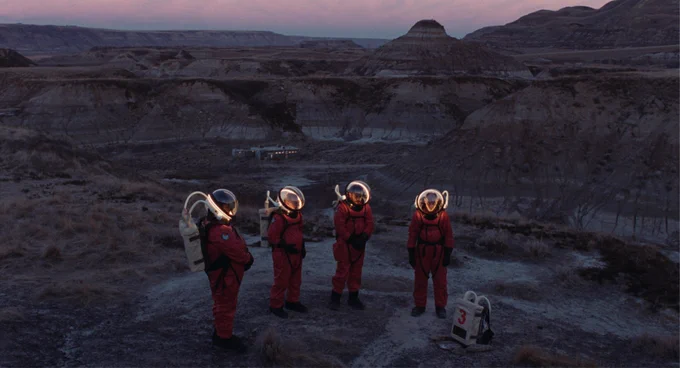 Four people, wearing orange astronaut suits with illuminated helmets, stand around a backpack with the number 3 on it. They are in an arid canyon with a building in the background in the film Viking.
