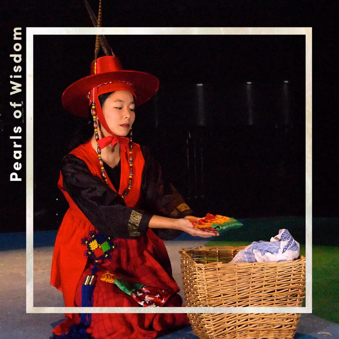 Sara Ochs holding a piece of folded cloth and kneeling on the floor in traditional attire. She looks very elegant, with a red satin hat with two pheasant feathers in it and a beaded tie. 